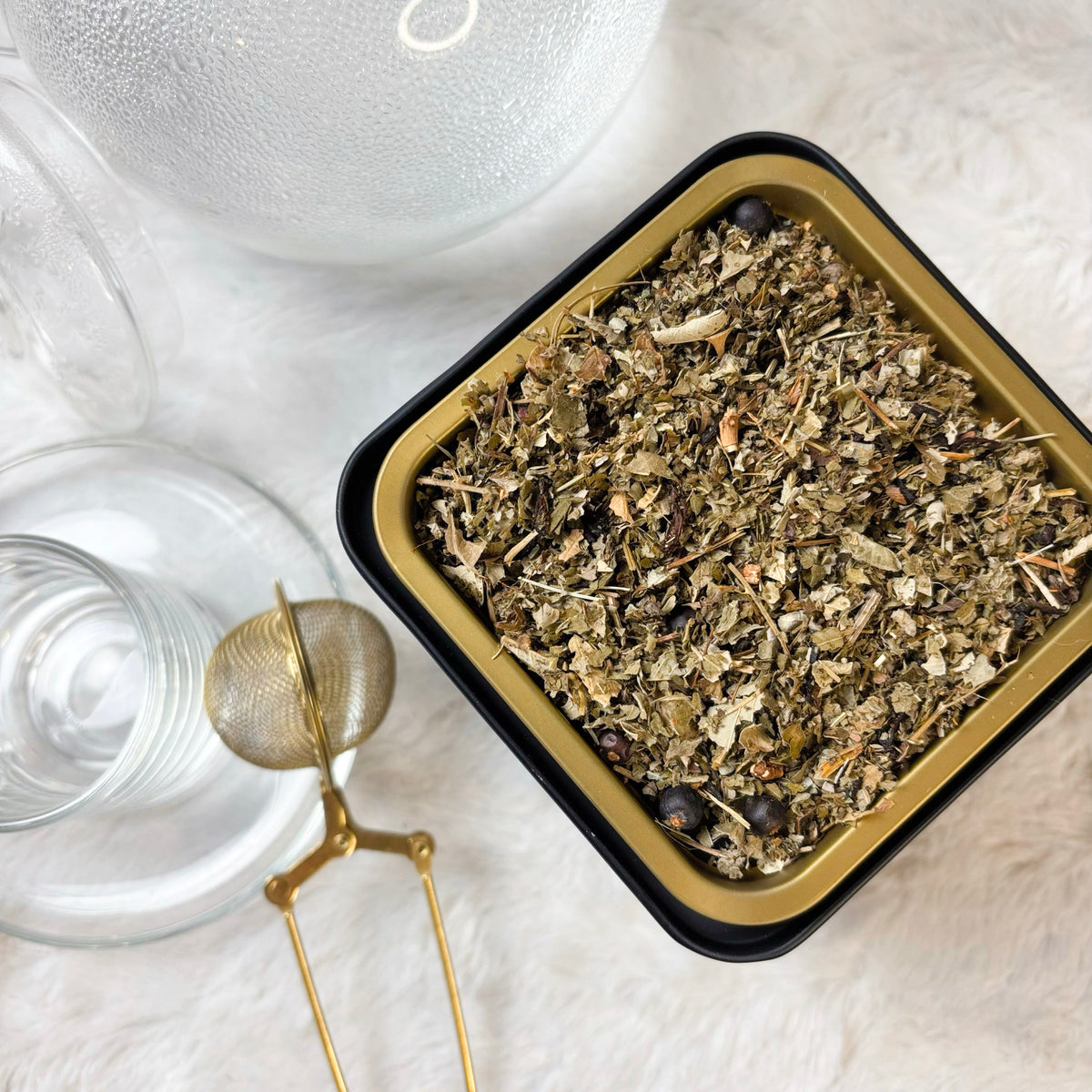 Herbal tea leaves in a black container with two glass cups and a gold tea spoon on a light surface.