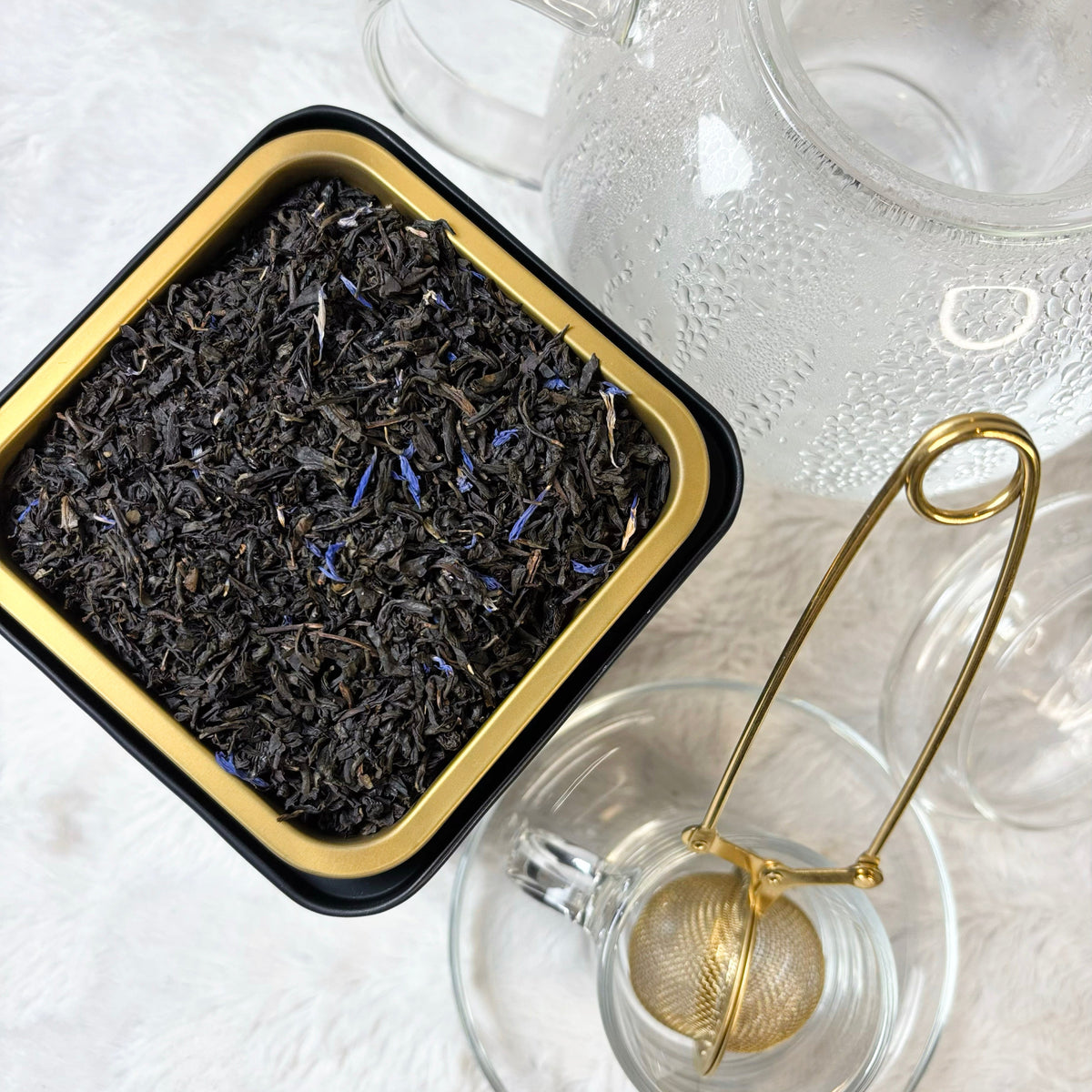 Tea leaves in a black and gold tin with a glass teapot and tea ball on a white surface