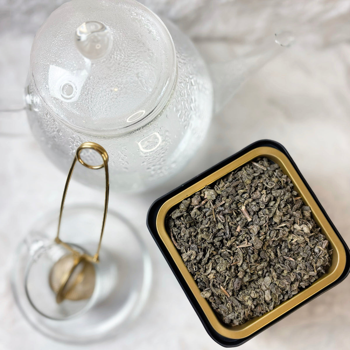 Clear glass jar with a lid and gold spoon next to a small black container with dried herbs on a white surface.