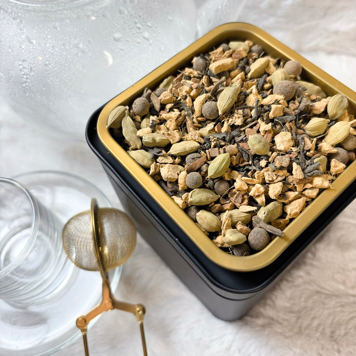 Herbal tea mix in a black and gold tin with glasses and a tea infuser on a white surface.
