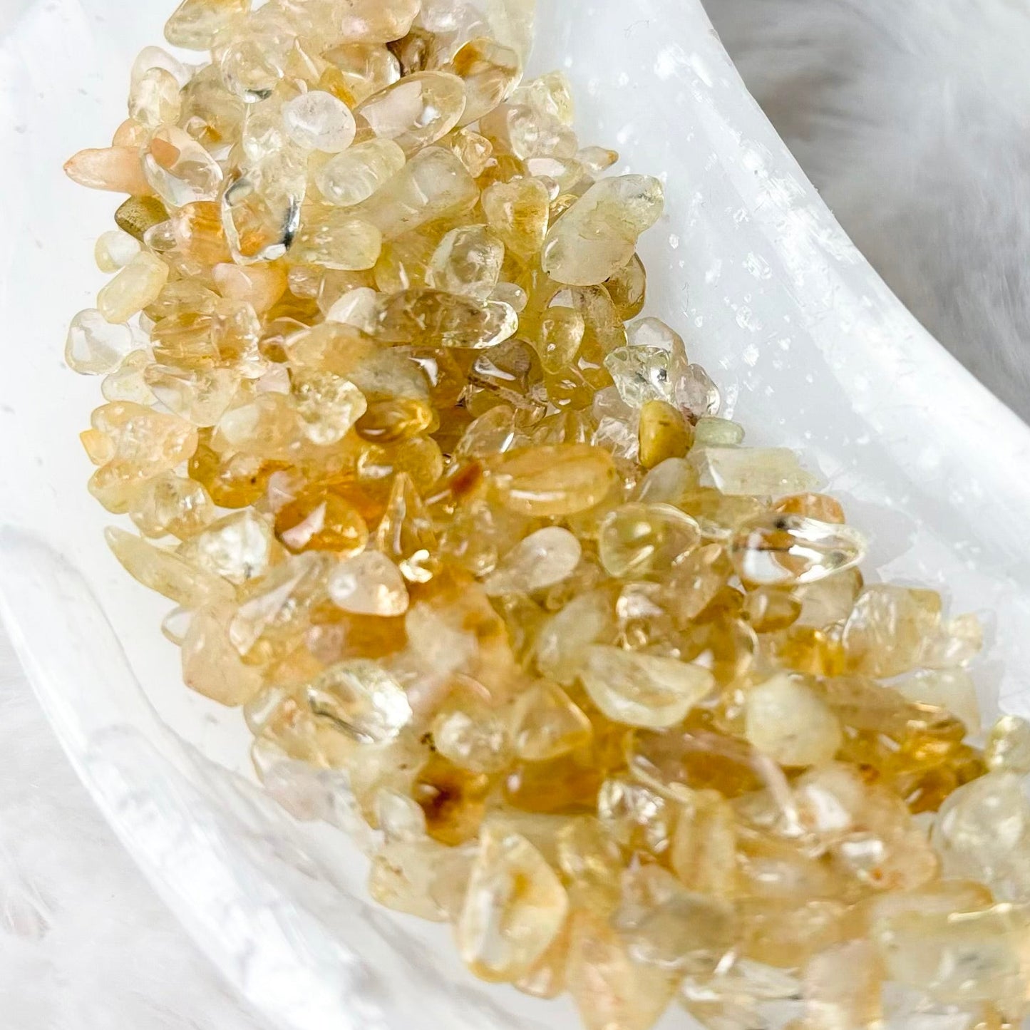 A cluster of small, translucent golden-yellow crystals arranged in a white crescent-shaped dish on a soft white faux fur surface.