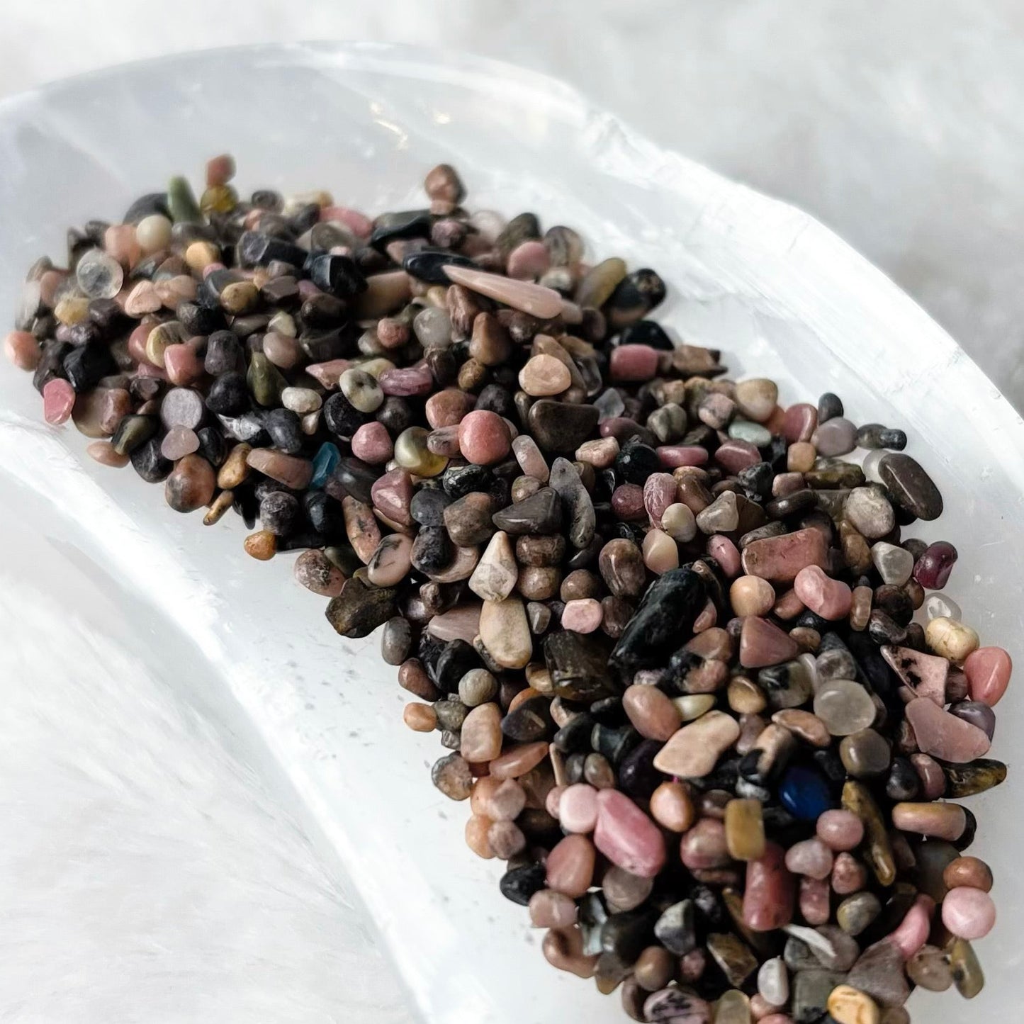 A small pile of multicolored pebbles or decorative stones sits in a white ceramic dish on a soft white faux-fur surface.