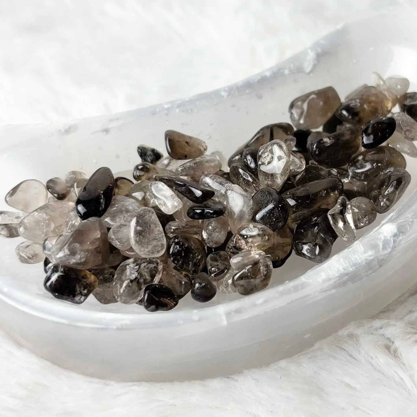 A small white crescent-shaped dish filled with polished smoky gray and black gemstone chips, placed on a soft white faux-fur surface.