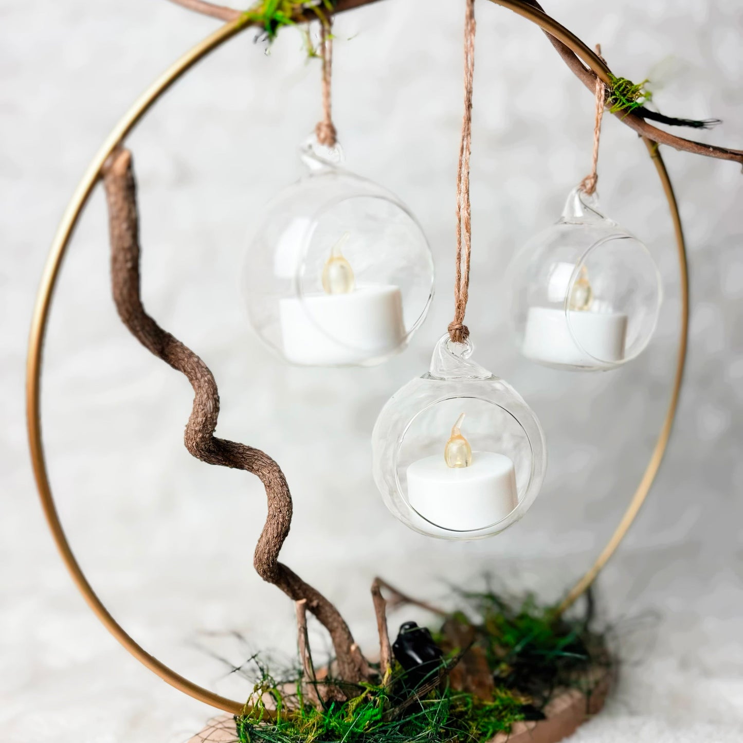 A decorative hanging candle display made from a natural branch ring and wooden base, with three clear glass hanging spheres containing white tea light candles, set against a soft white textured background.