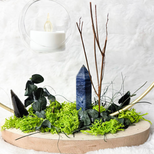 A decorative natural stone arrangement on a round wooden tray, featuring a blue lapis lazuli crystal point, black agate pieces, green moss, eucalyptus leaves, and bare twigs, set against a white faux fur background with hanging glass candle holders.