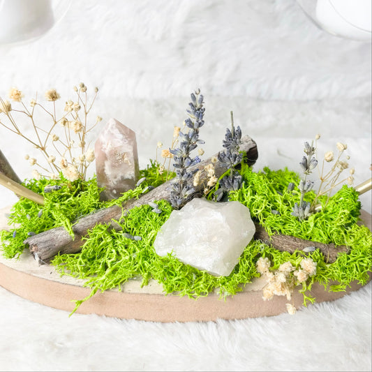 A decorative oval wooden tray filled with bright green moss, dried branches, small pale flowers, and natural stones, styled on a white faux-fur surface with blurred glass candle holders in the background.