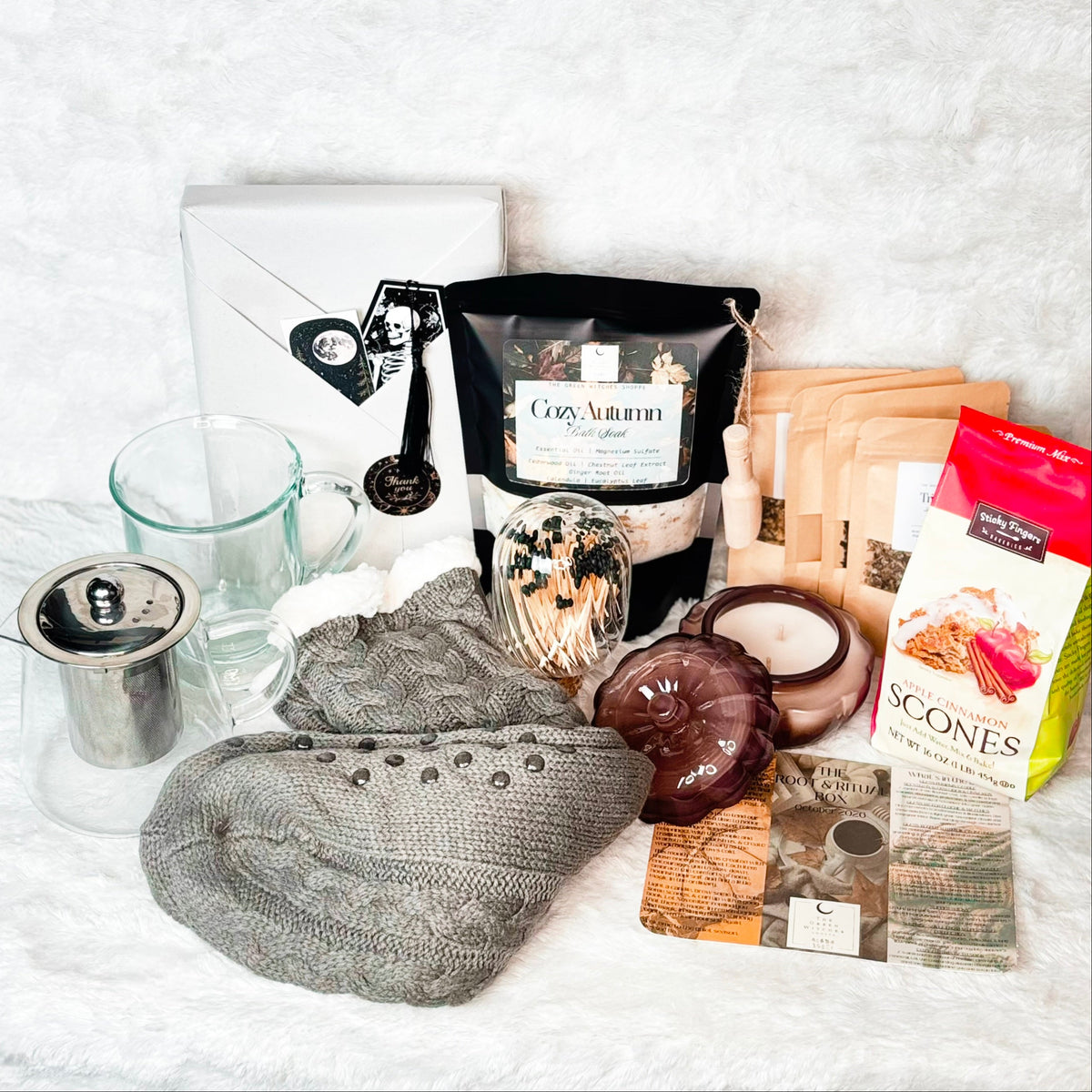 A cozy holiday gift set arranged on a white faux fur background, featuring gray cable-knit slipper socks, a glass mug and teapot with metal infuser, a bag of Cozy Autumn Bath Soak, a box of gourmet scone mix, a candle, and other warm seasonal accessories.