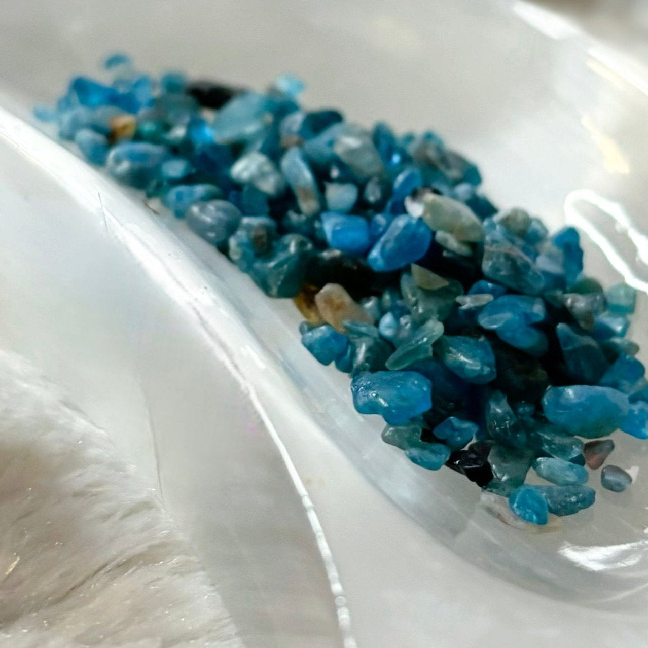 Small bottle labeled 'Blue Apatite' with blue stones on a white dish against a blurred background of similar bottles.