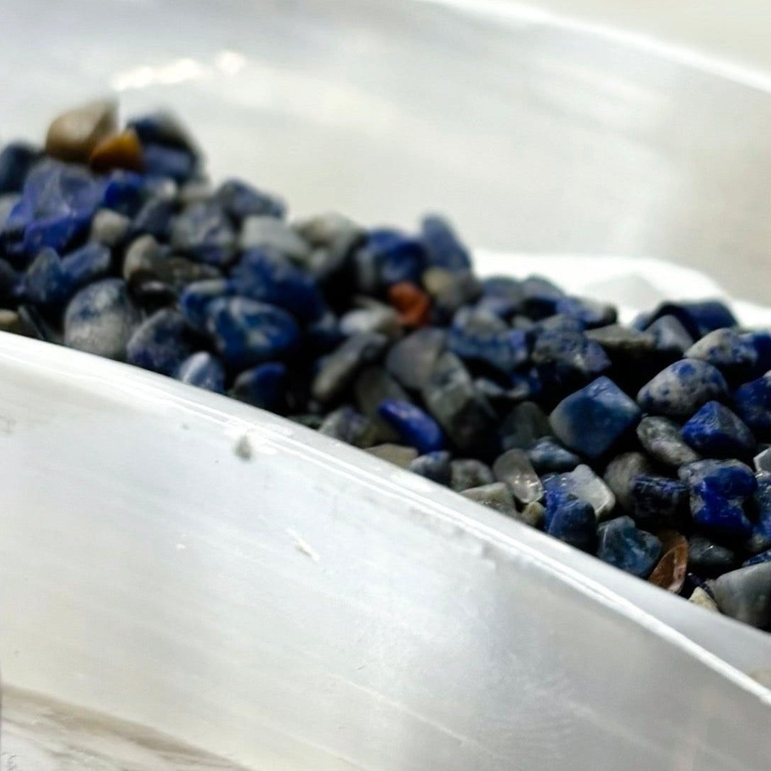 Small glass bottle labeled 'Lapis Lazuli' with a white dish containing blue stones on a white fur surface.