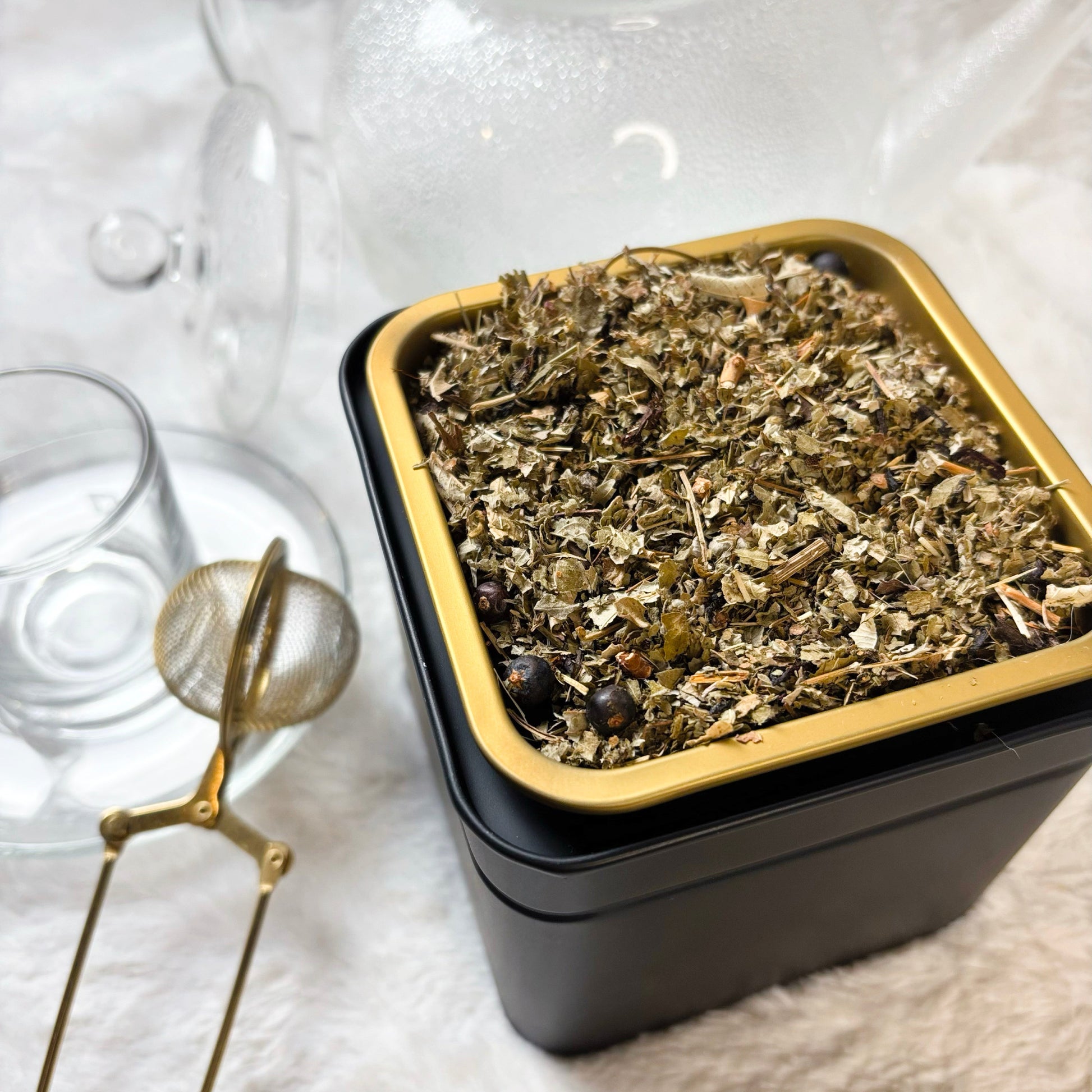 Black container with gold lid filled with tea leaves on a white surface with a teapot and glass in the background.