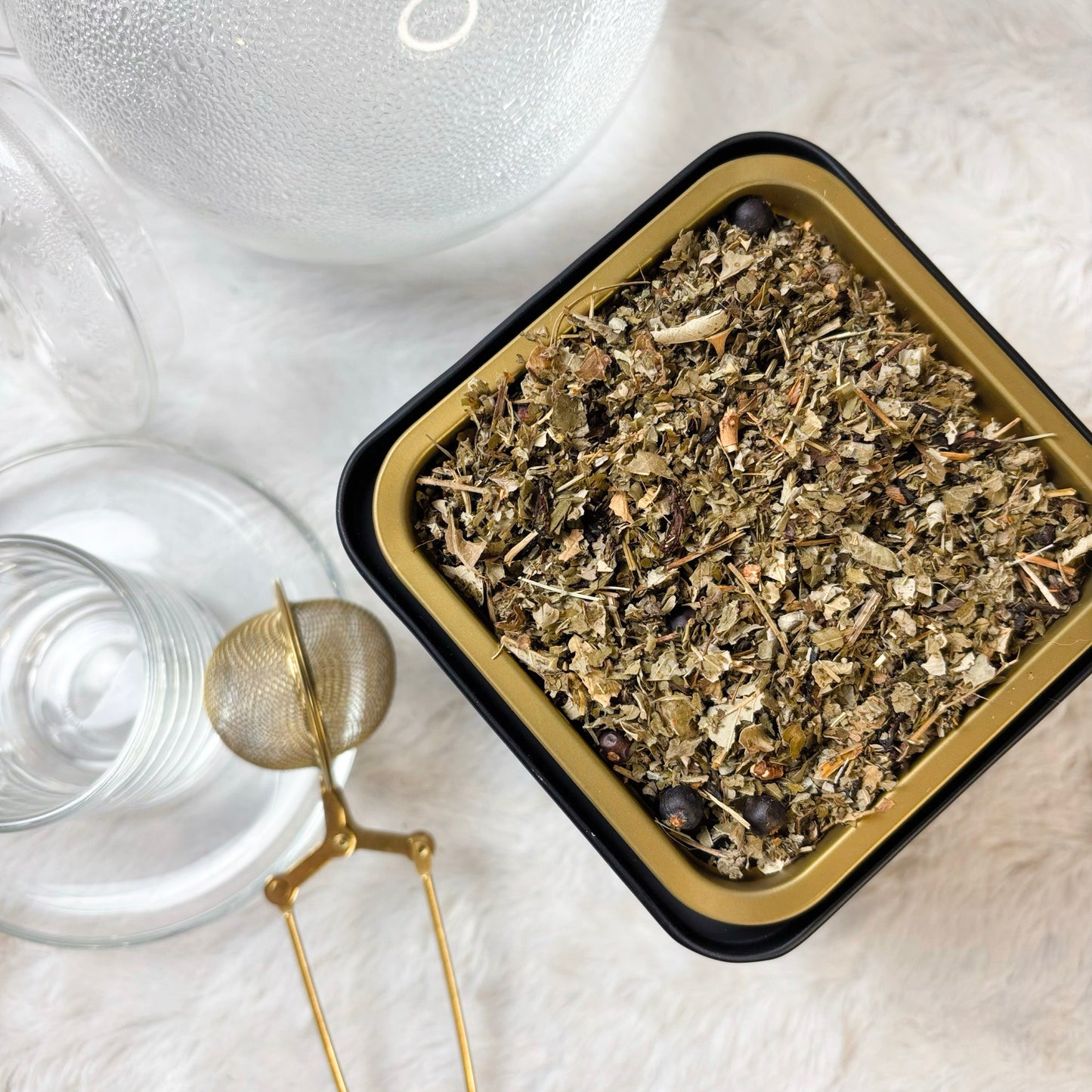 Herbal tea leaves in a black container with two glass cups and a gold tea spoon on a light surface.