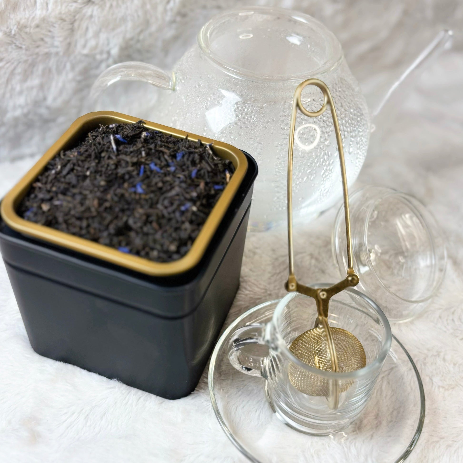 Black container with dried herbs, glass teacup, and strainer on a textured surface
