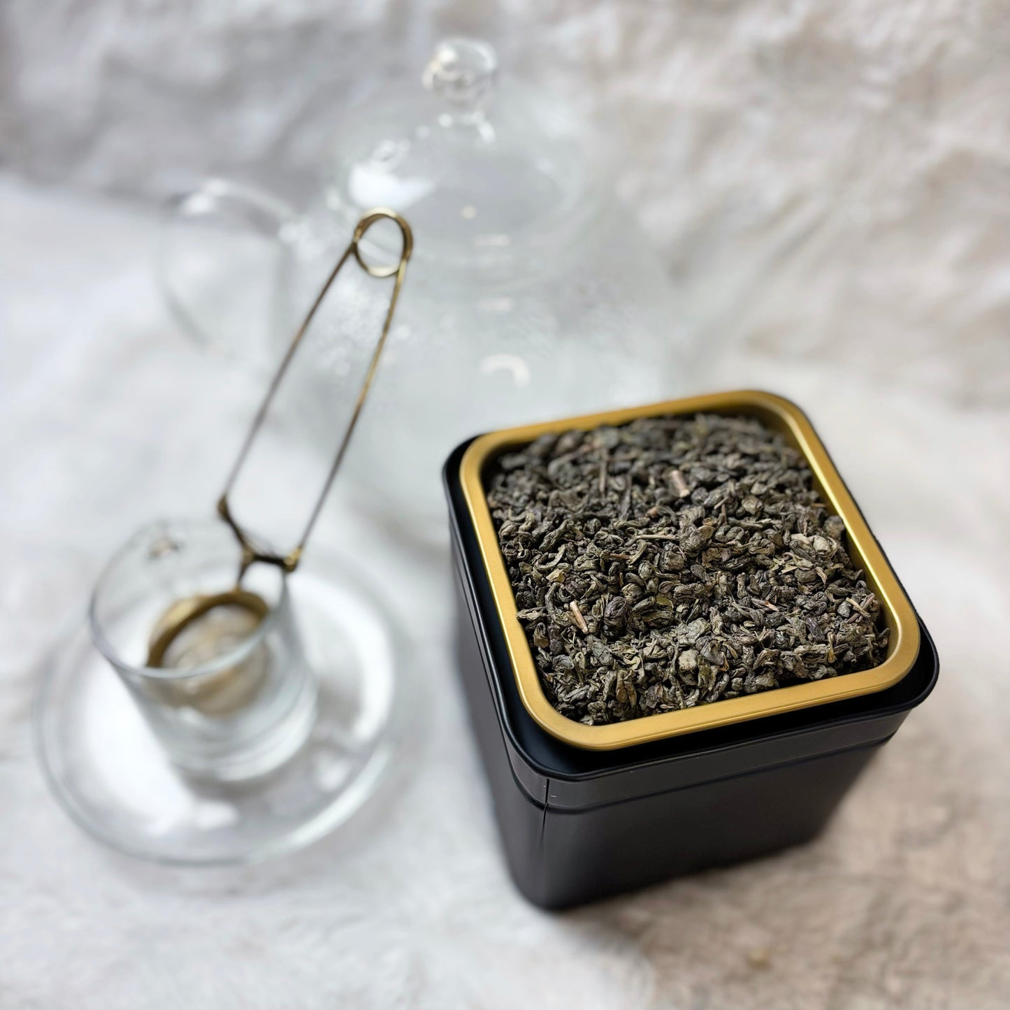 Square black container with gold rim filled with tea leaves next to a glass teacup on a white textured surface.