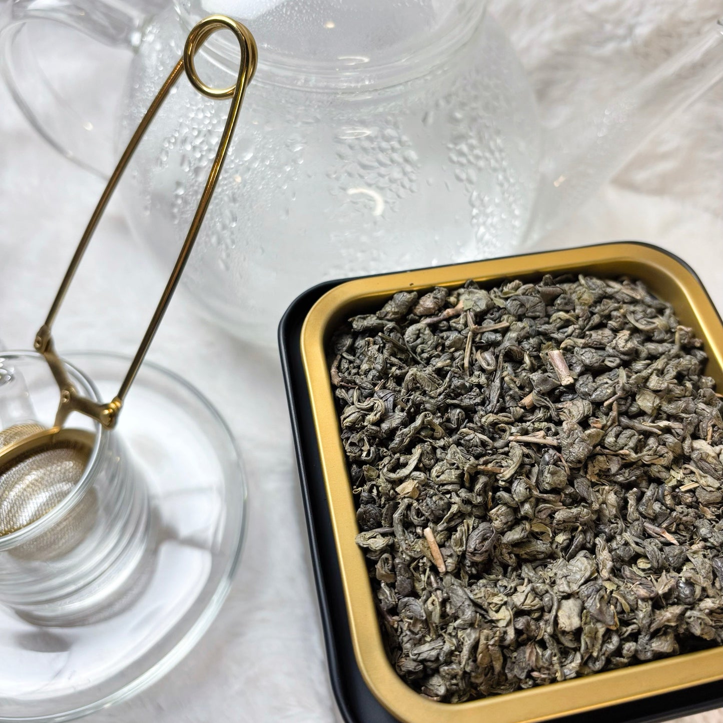 Tea leaves in a black and gold tin with a glass teapot and strainer on a white surface.
