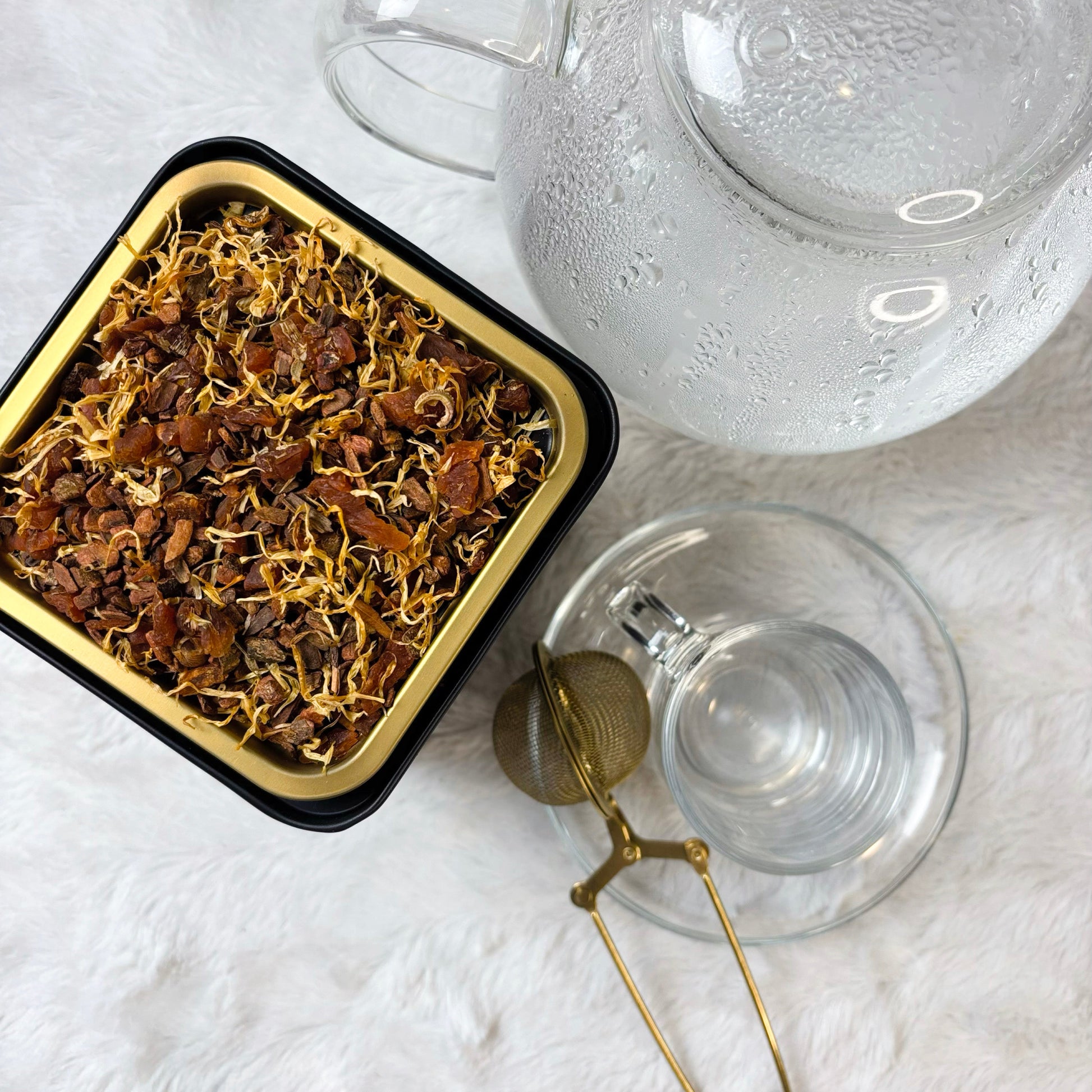 Tea leaves in a black container with a clear teapot on a white surface