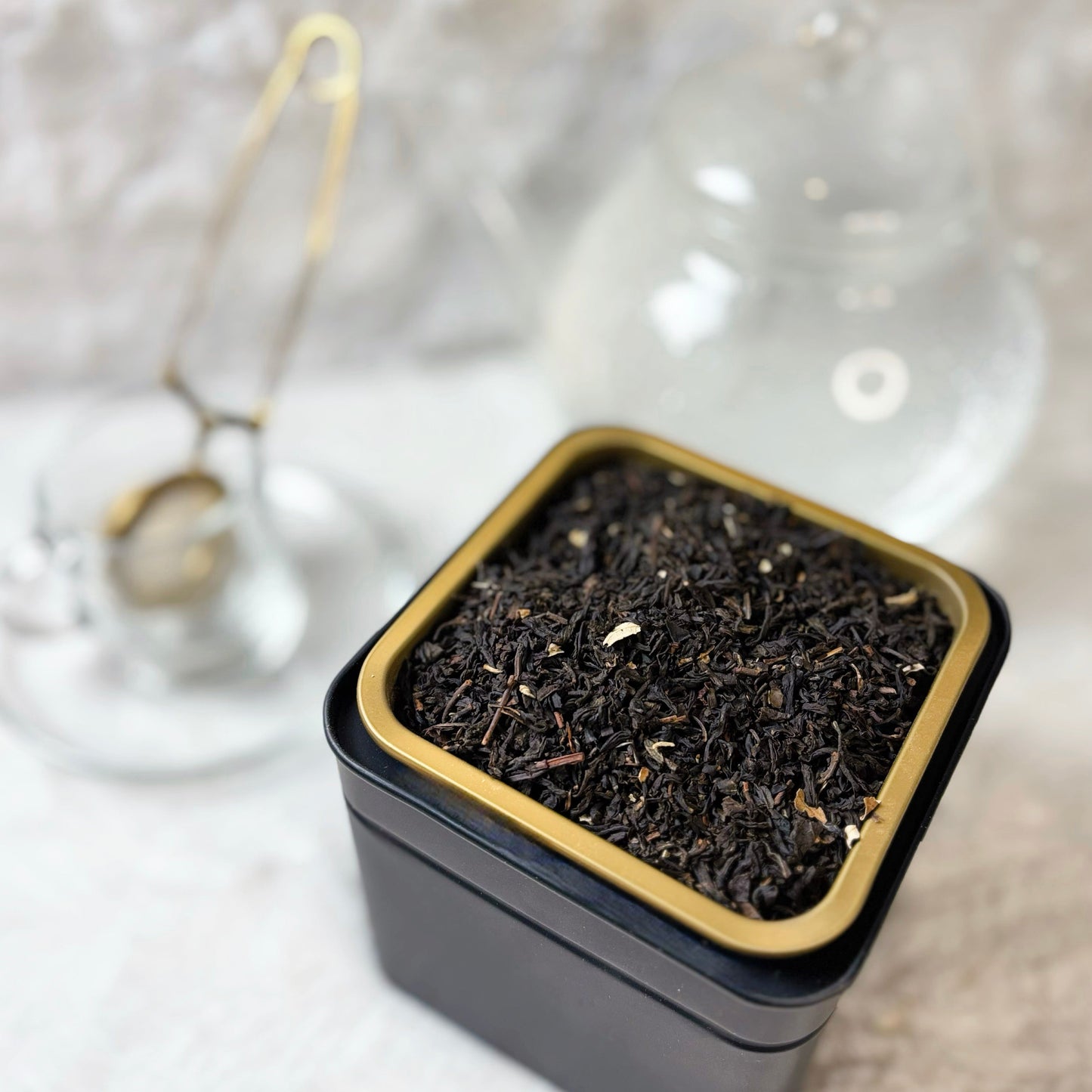 Black tea leaves in a small square tin with gold rim on a textured white surface.
