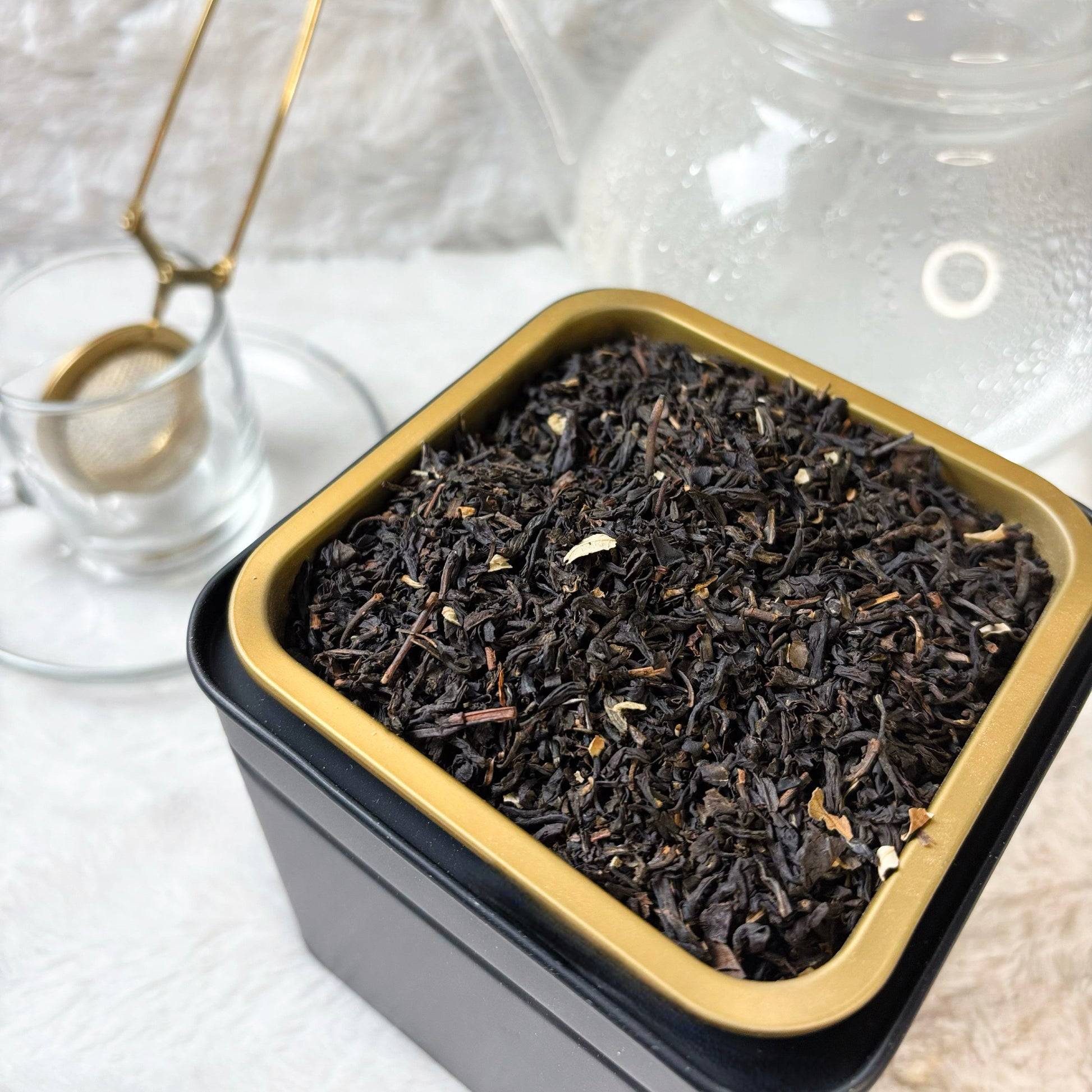 Black tea leaves in a square metal tin with a gold rim on a light background