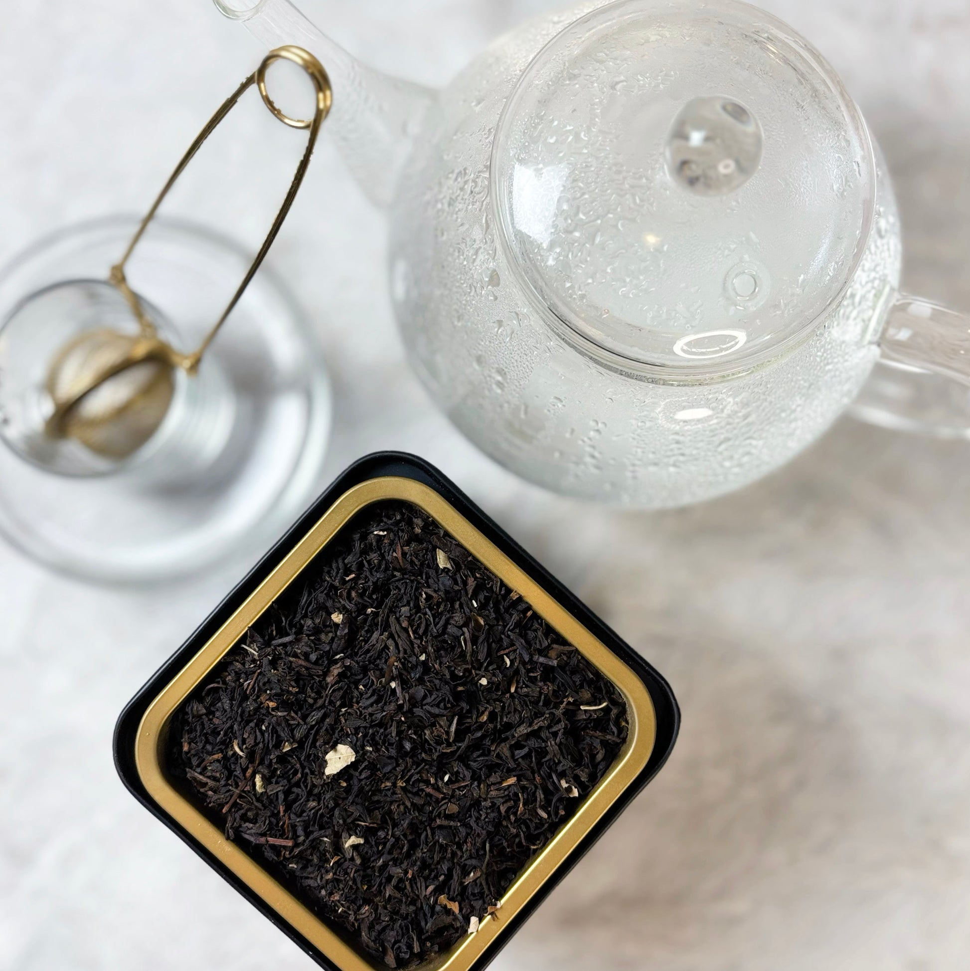 Clear glass teapot with a lid, gold tea infuser, and container of black tea leaves on a white surface.