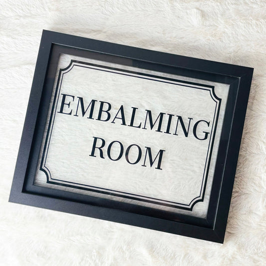 Framed sign with 'EMBALMING ROOM' text on a textured white background