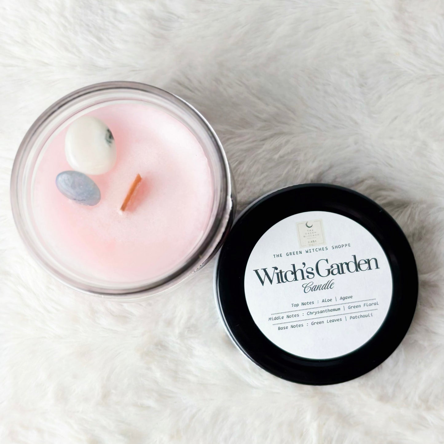 Pink candle in a clear container with a black lid labeled 'Witch's Garden' on a white fluffy surface.