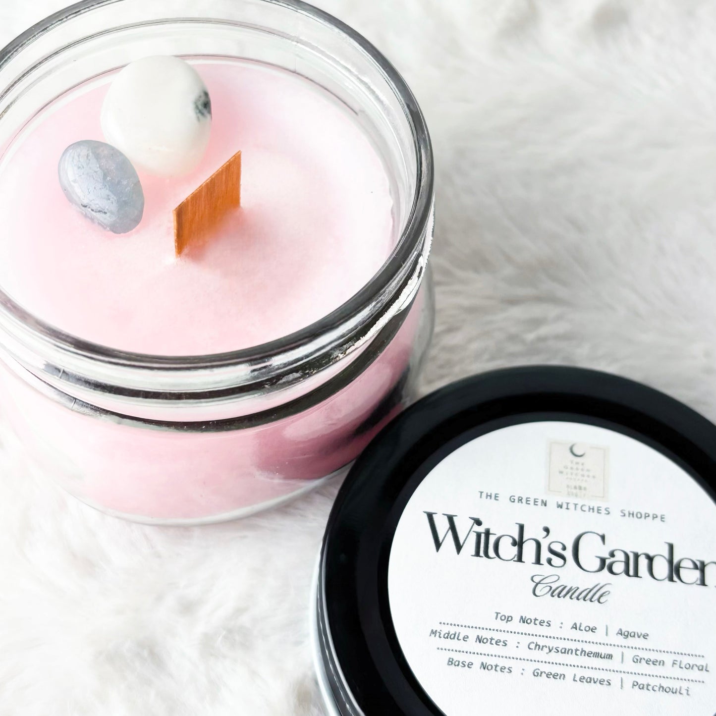 Pink candle in a glass jar with a black lid labeled 'Witch's Garden' on a white fabric background.