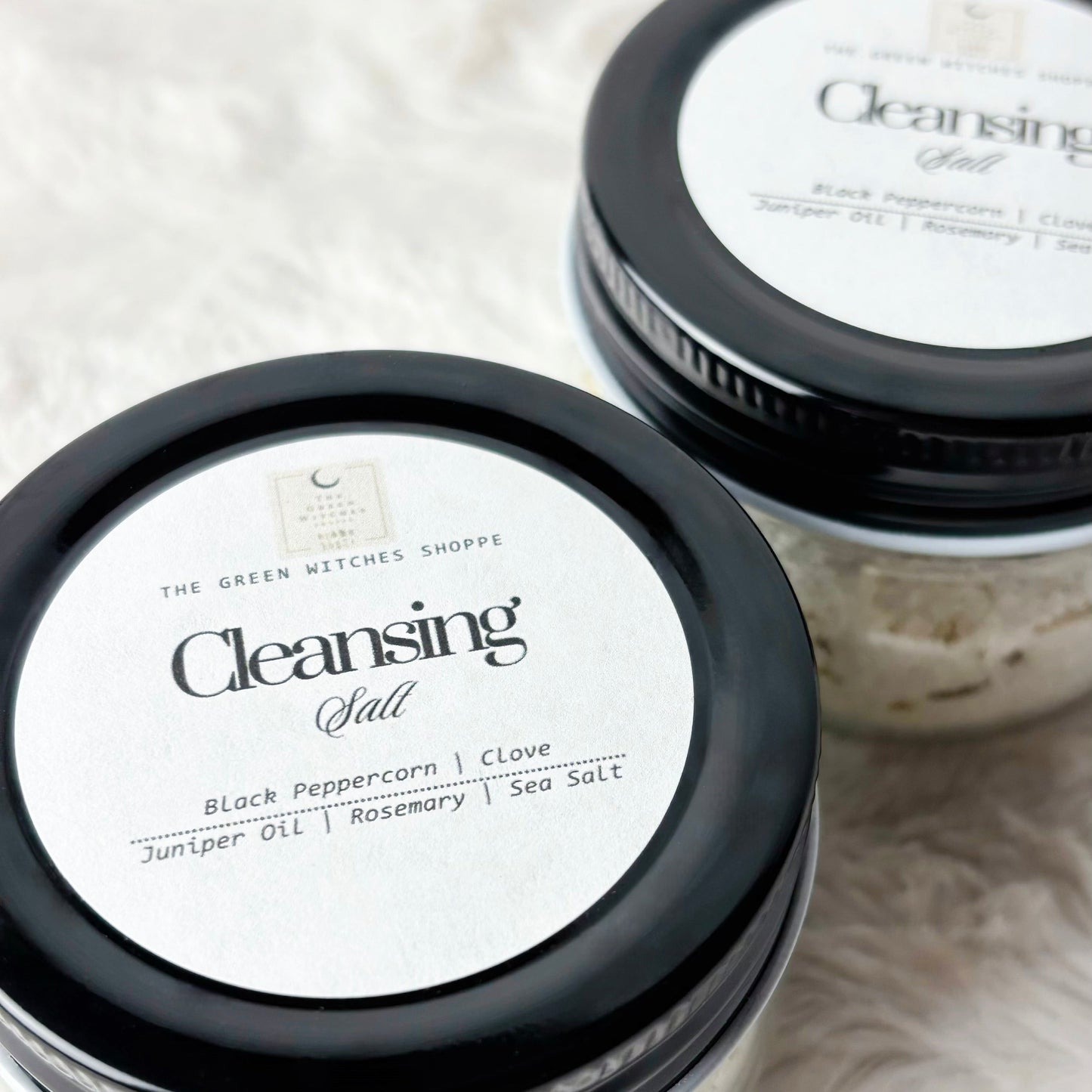 Two jars of 'Cleansing Salt' with black lids on a textured white surface.