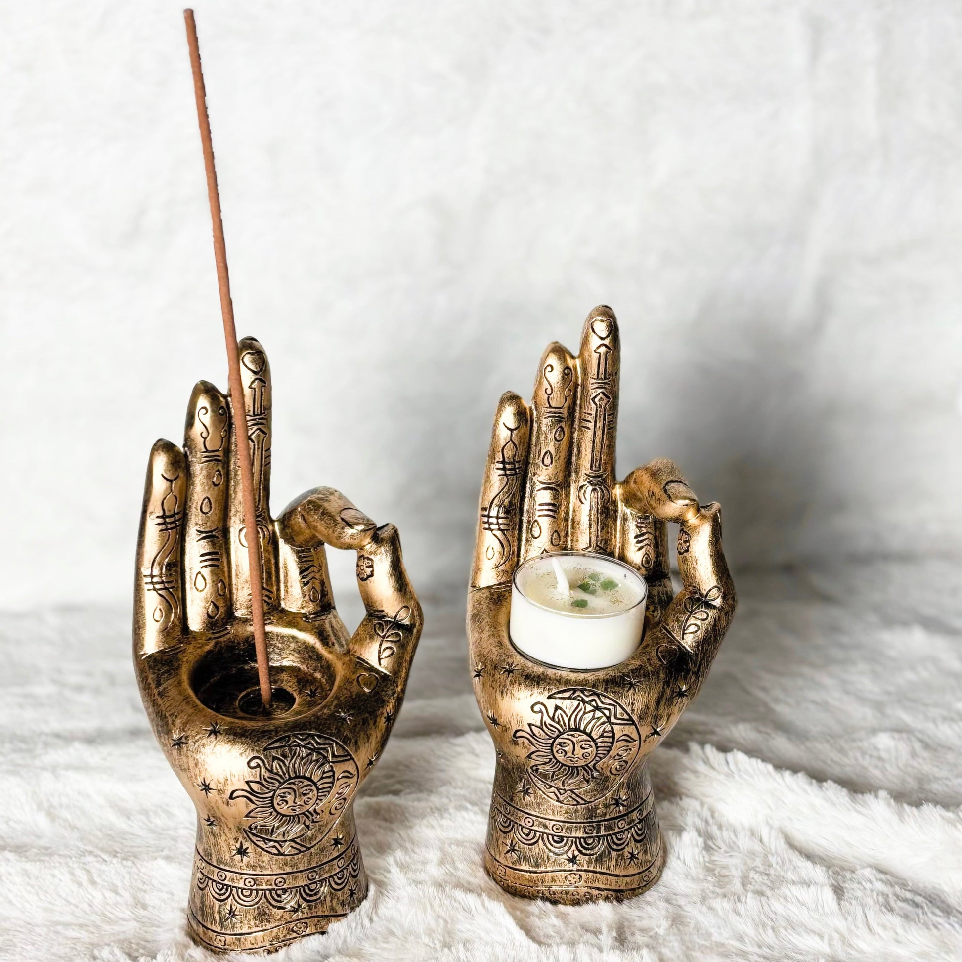 Bronze hand-shaped candle holders with one holding a candle and the other an incense stick on a white fabric background.