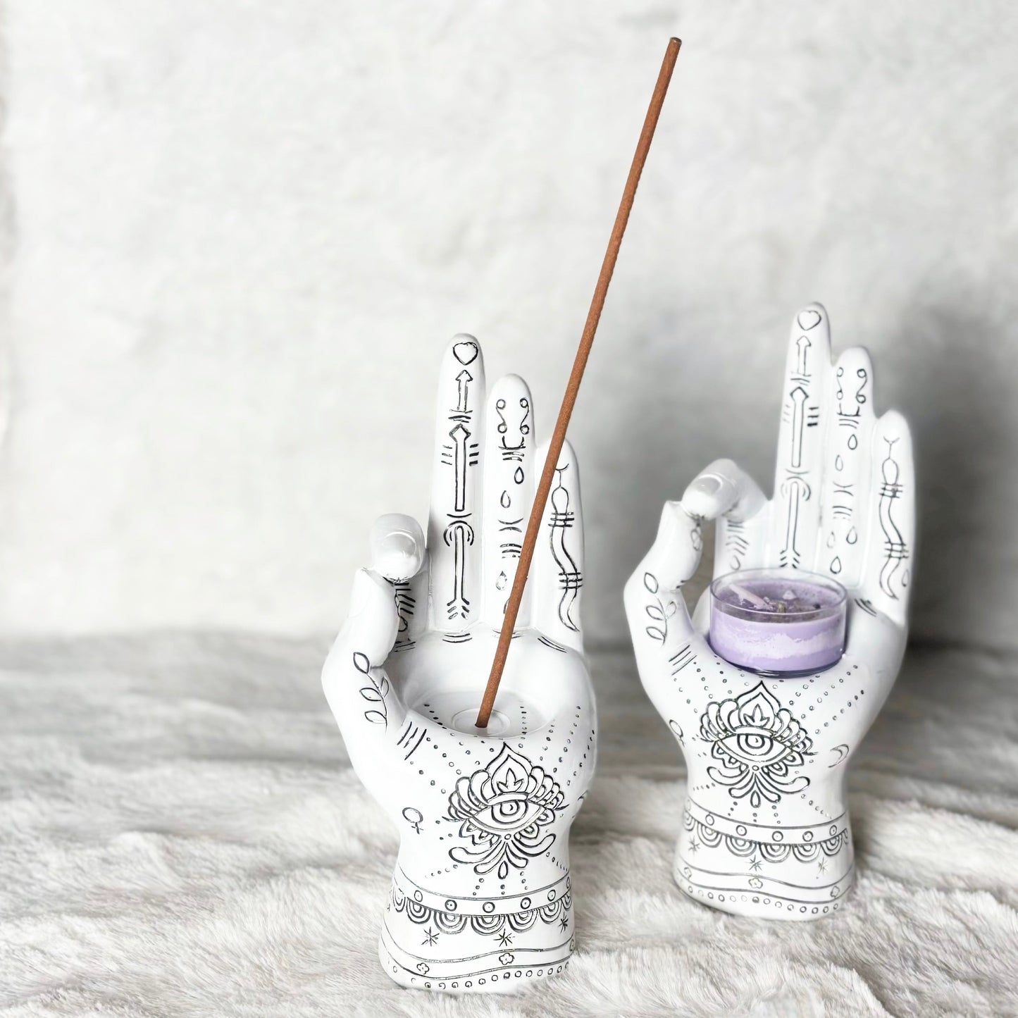 Decorative hands holding a candle and an incense stick on a textured surface.