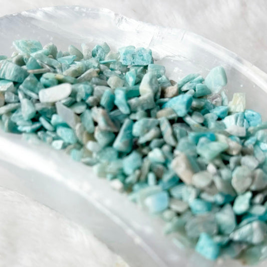 Light blue and white raw amethyst chip bath salts resting in a clear glass bowl on a white faux fur surface.
