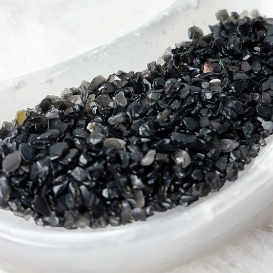 Coarse black volcanic gravel or crushed charcoal stones piled in a small white ceramic dish on a white faux fur background.