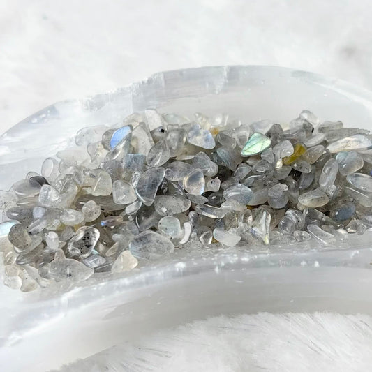 A crescent-shaped clear resin tray filled with small gray and iridescent labradorite chips, resting on a white faux fur surface.