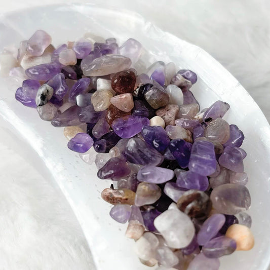 Purple and lavender amethyst tumbled stone chips arranged in a white crescent-shaped dish on a soft white faux fur background.
