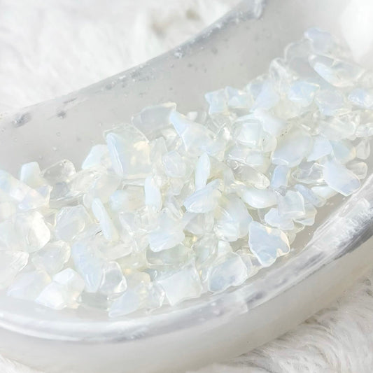 White iridescent crystal chips arranged in a small translucent crescent-shaped dish on a soft white faux-fur surface.