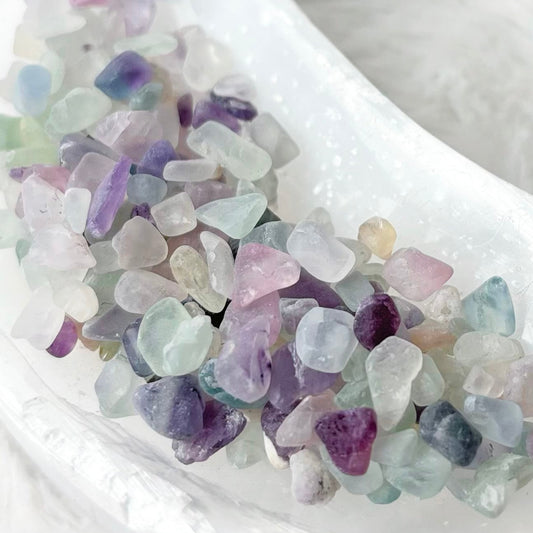 A small white crescent-shaped dish filled with polished tumbled gemstones in pastel shades of purple, blue, green, and pink, set on a soft white faux fur surface.