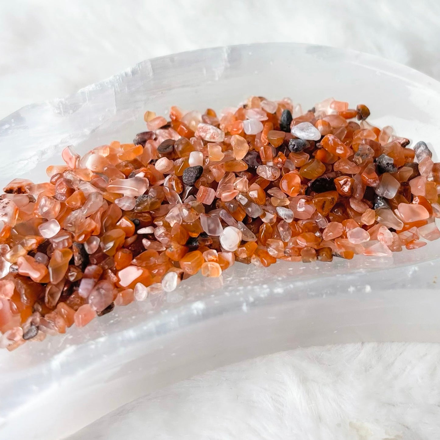 A curved clear resin tray holding a bed of small peach, pink, and brown crystal chips, photographed on a soft white faux-fur surface.