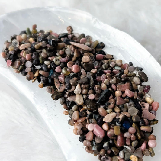 A small pile of multicolored pebbles or decorative stones sits in a white ceramic dish on a soft white faux-fur surface.