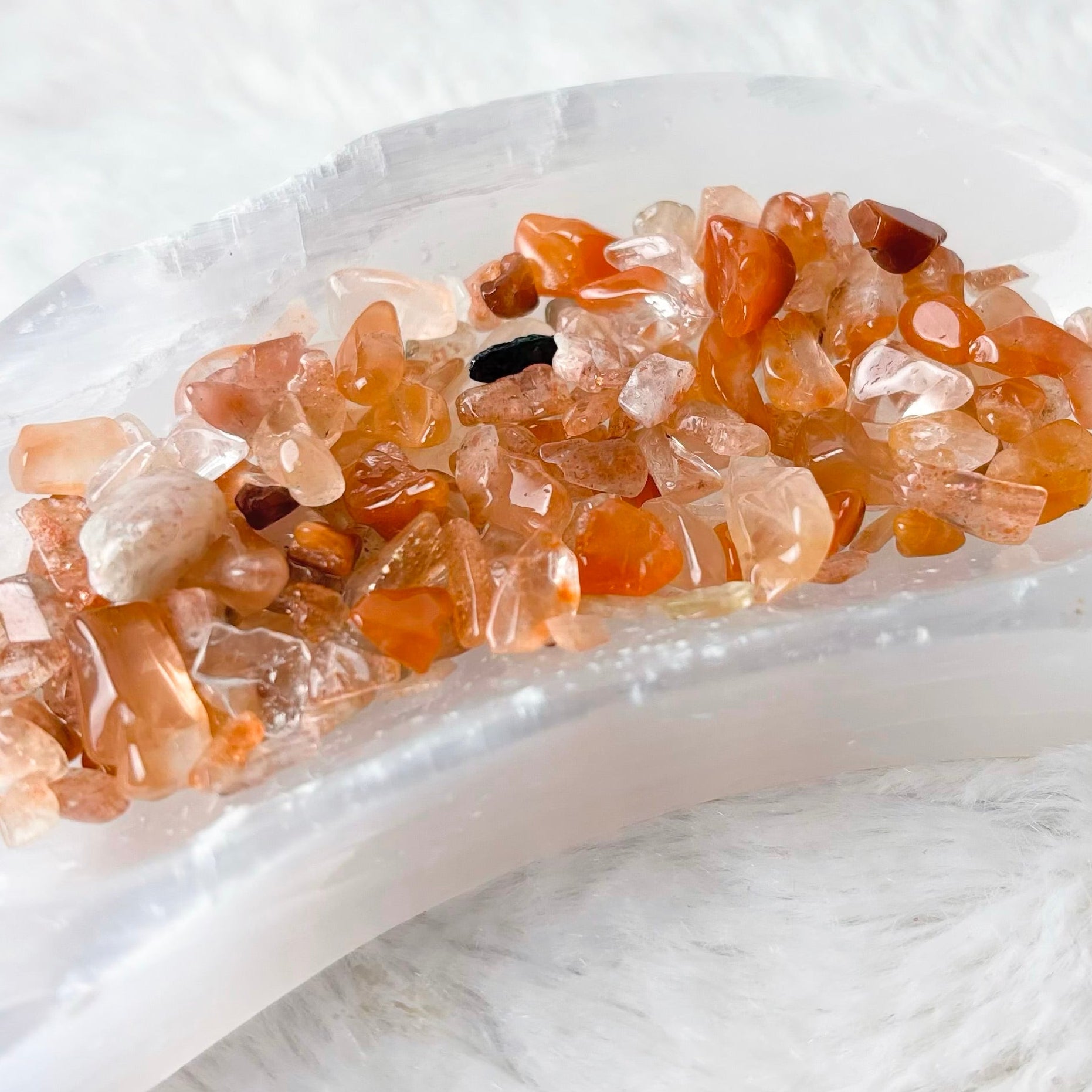 A crescent-shaped white crystal cluster holder or tray filled with small orange, peach, and amber crystals, resting on a white faux fur surface.