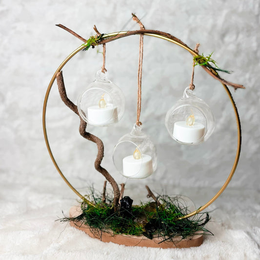 A decorative hanging light fixture made from a circular gold metal hoop, natural branches, moss, and a small wooden base, with three clear glass domes holding white tea light candles, photographed against a soft white textured backdrop.