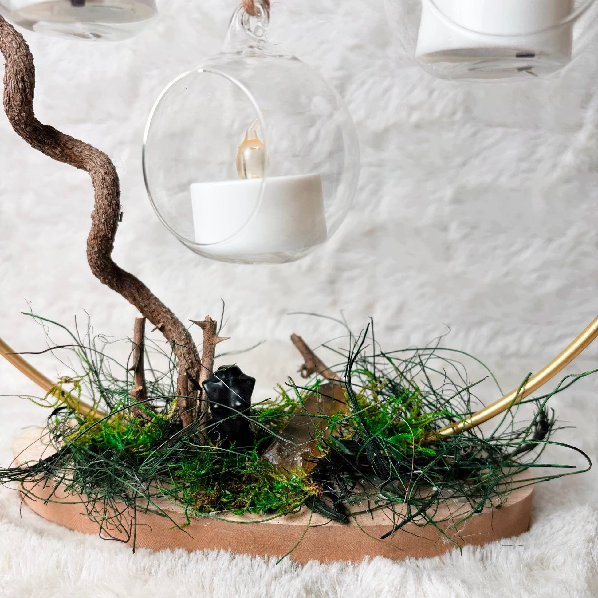 A decorative tabletop arrangement on a round wooden base covered with green moss, thin black twigs, and gold-toned curved rods, with a gnarled brown branch rising upward. Three clear glass hanging lanterns with white tea light candles are suspended above against a soft white textured background.