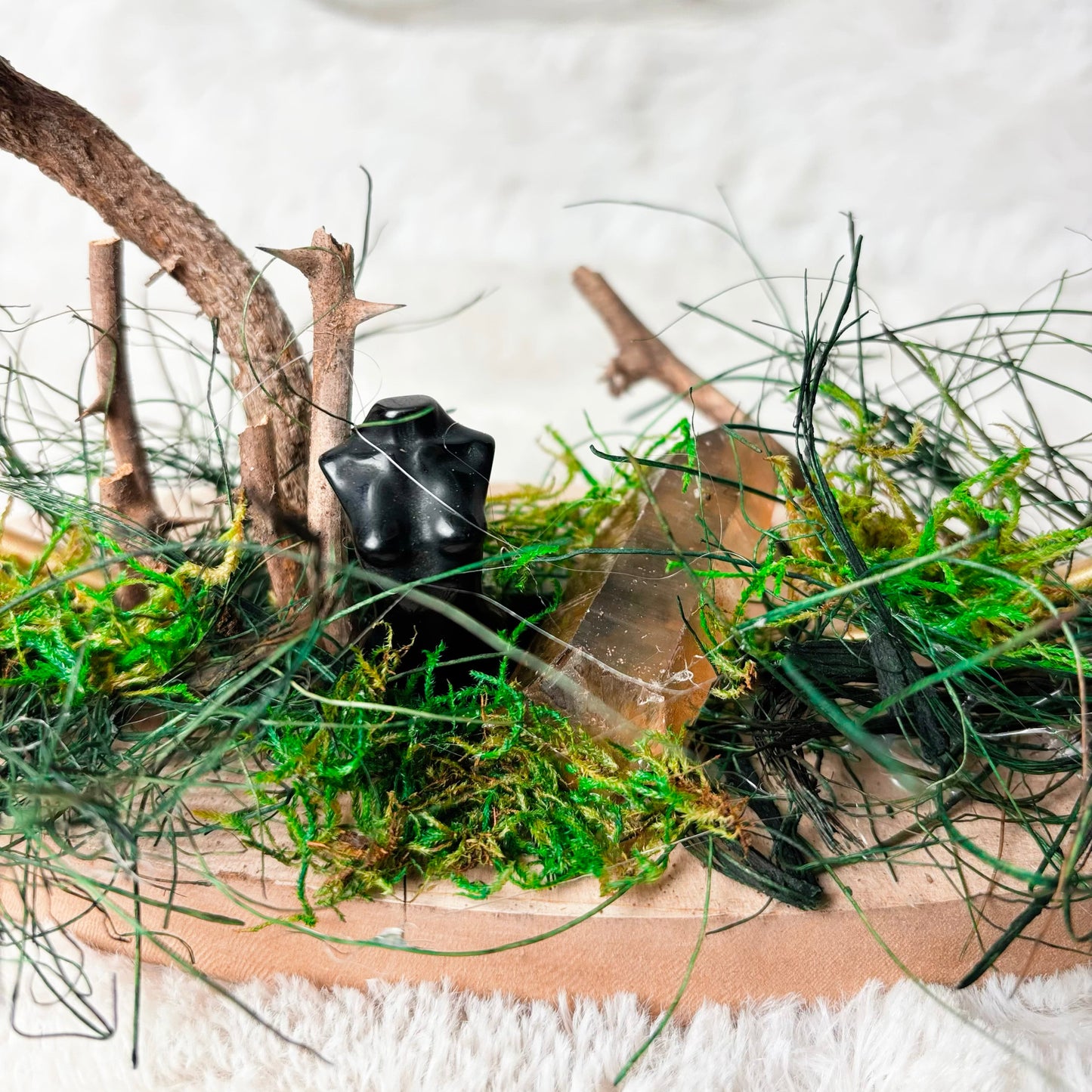 A miniature fairy garden scene on a wooden tray, featuring green moss, artificial grass, small twigs, and a small black frog figurine, set against a soft white textured background.