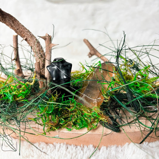 A miniature fairy garden scene on a wooden tray, featuring green moss, artificial grass, small twigs, and a small black frog figurine, set against a soft white textured background.