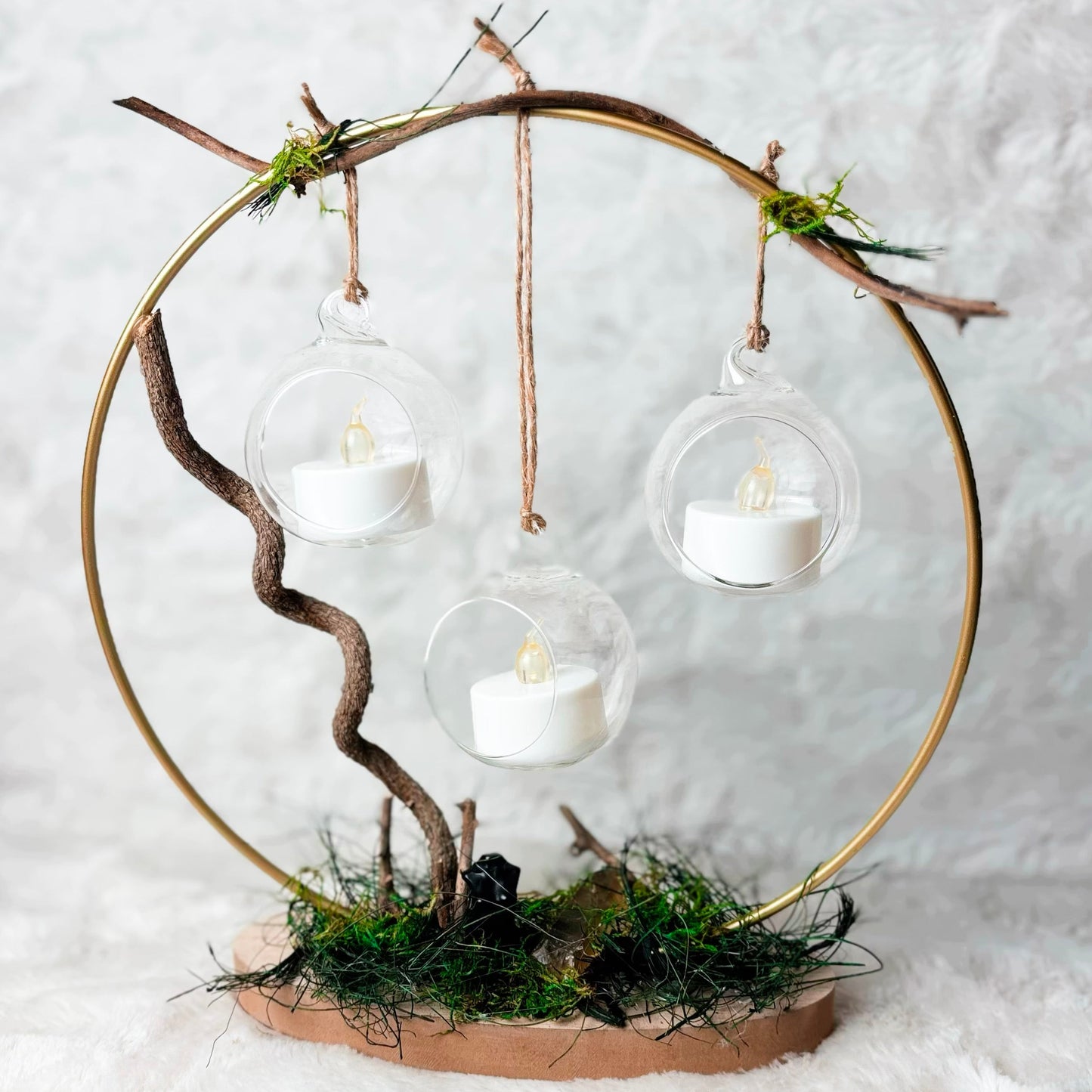 A rustic decorative hanging circle centerpiece made from a gold metal hoop, twine, small branches, moss, and a brown base, with three clear glass globes containing white tea light candles, set against a white textured background.