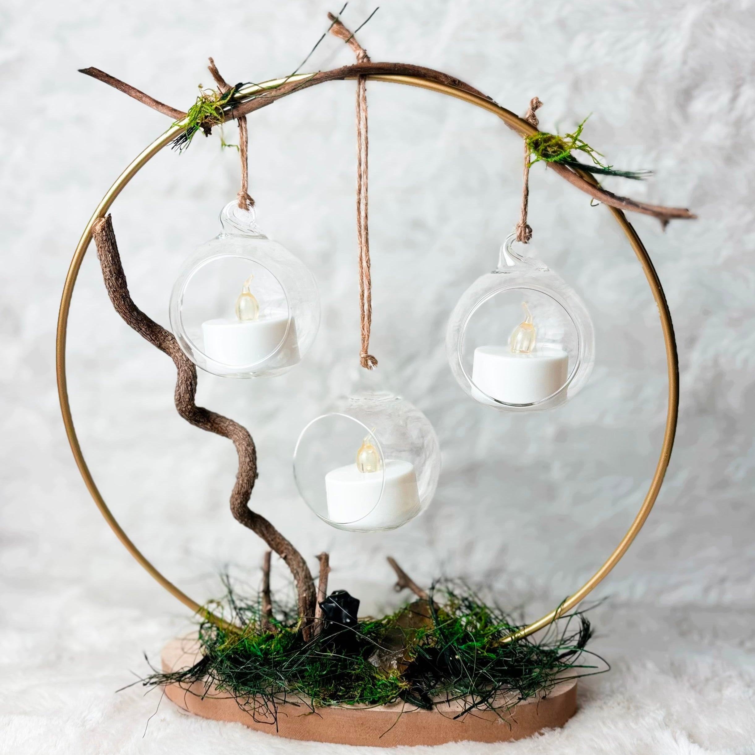 A rustic decorative hanging circle centerpiece made from a gold metal hoop, twine, small branches, moss, and a brown base, with three clear glass globes containing white tea light candles, set against a white textured background.