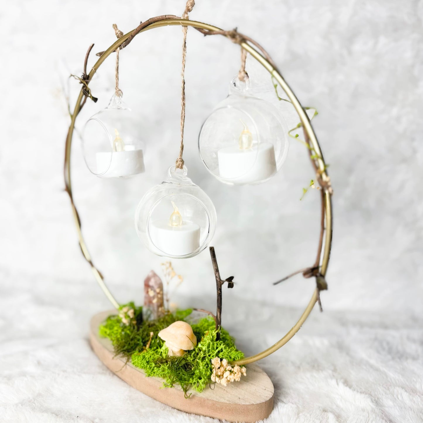 A decorative hanging lantern arrangement with three clear glass globe cups suspended from a rustic circular metal frame, each holding a white tea light candle. The base is a natural wood slice decorated with green moss, small dried flowers, and tiny figurines, set against a soft white textured fabric background.