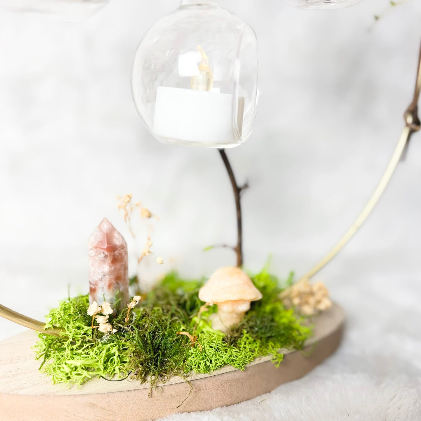 A decorative miniature fairy garden scene on a round wooden base, featuring green moss, a pink crystal, a small beige mushroom, dried flowers, and a clear glass dome hanging above with a white candle inside.