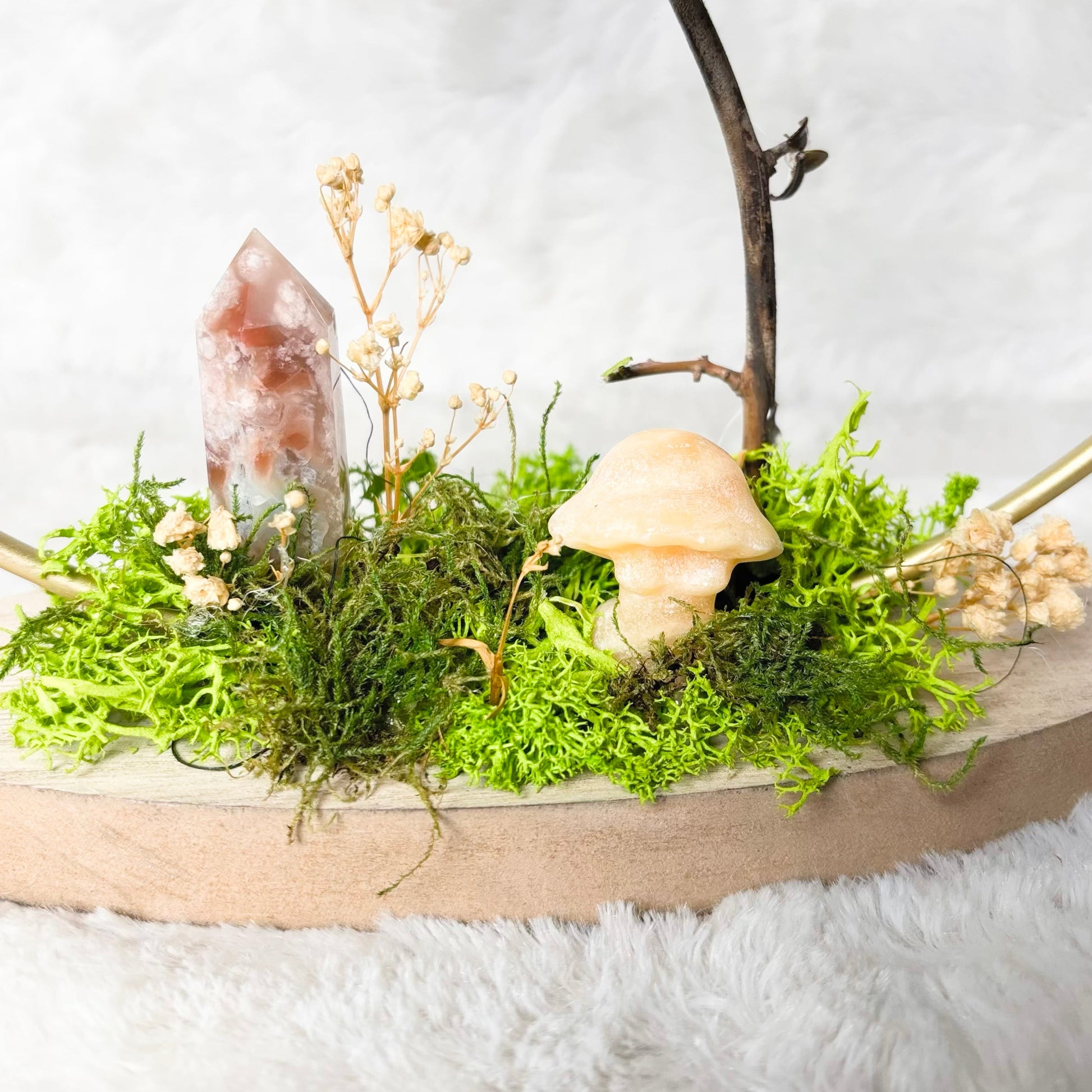 A decorative tabletop arrangement on a round wooden board featuring bright green moss, a clear crystal, a small beige mushroom figurine, dried flowers, a dark branch stem, and a white candle in a clear glass holder, set against a soft white textured background.
