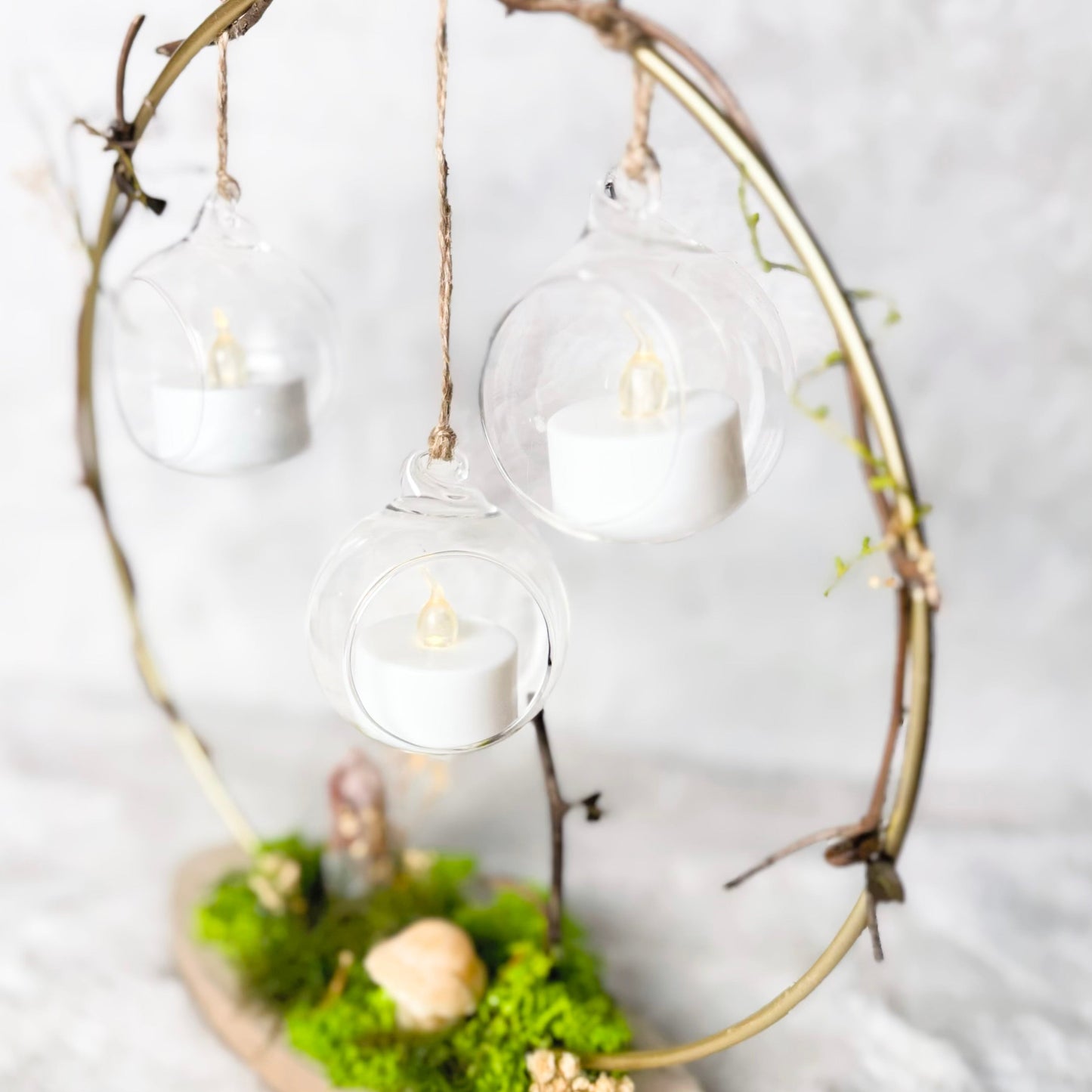 A decorative hanging candle display made from a natural twig circle frame, with three clear glass globes suspending white tea light candles, mounted on a small wooden base covered in green moss and dried botanicals, set against a soft white fabric background.