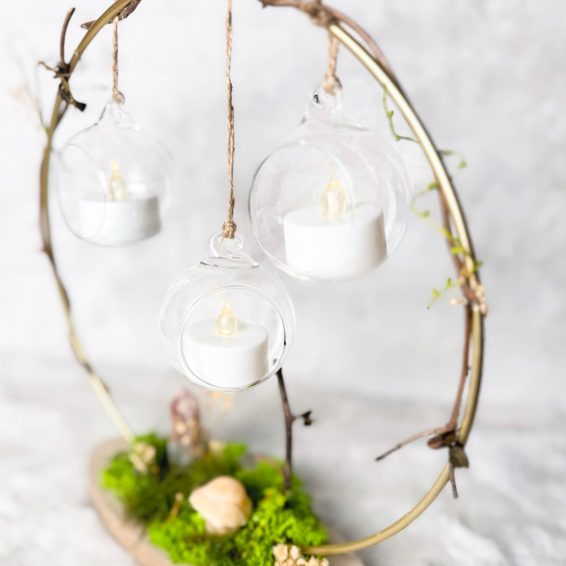 A decorative hanging candle display made from a natural twig circle frame, with three clear glass globes suspending white tea light candles, mounted on a small wooden base covered in green moss and dried botanicals, set against a soft white fabric background.