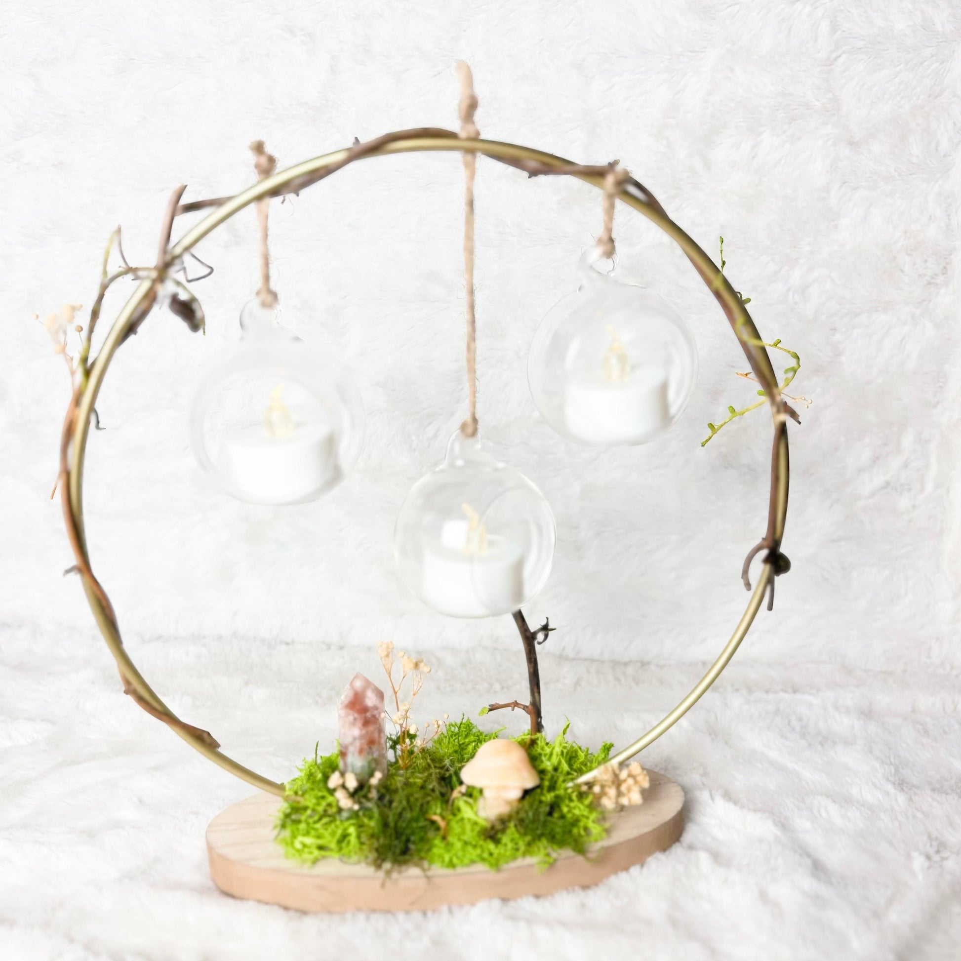 A rustic circular twig hanging lantern with three clear glass cups holding white tea light candles, mounted on a natural wood base with green moss and small dried flowers, photographed against a soft white textured backdrop.