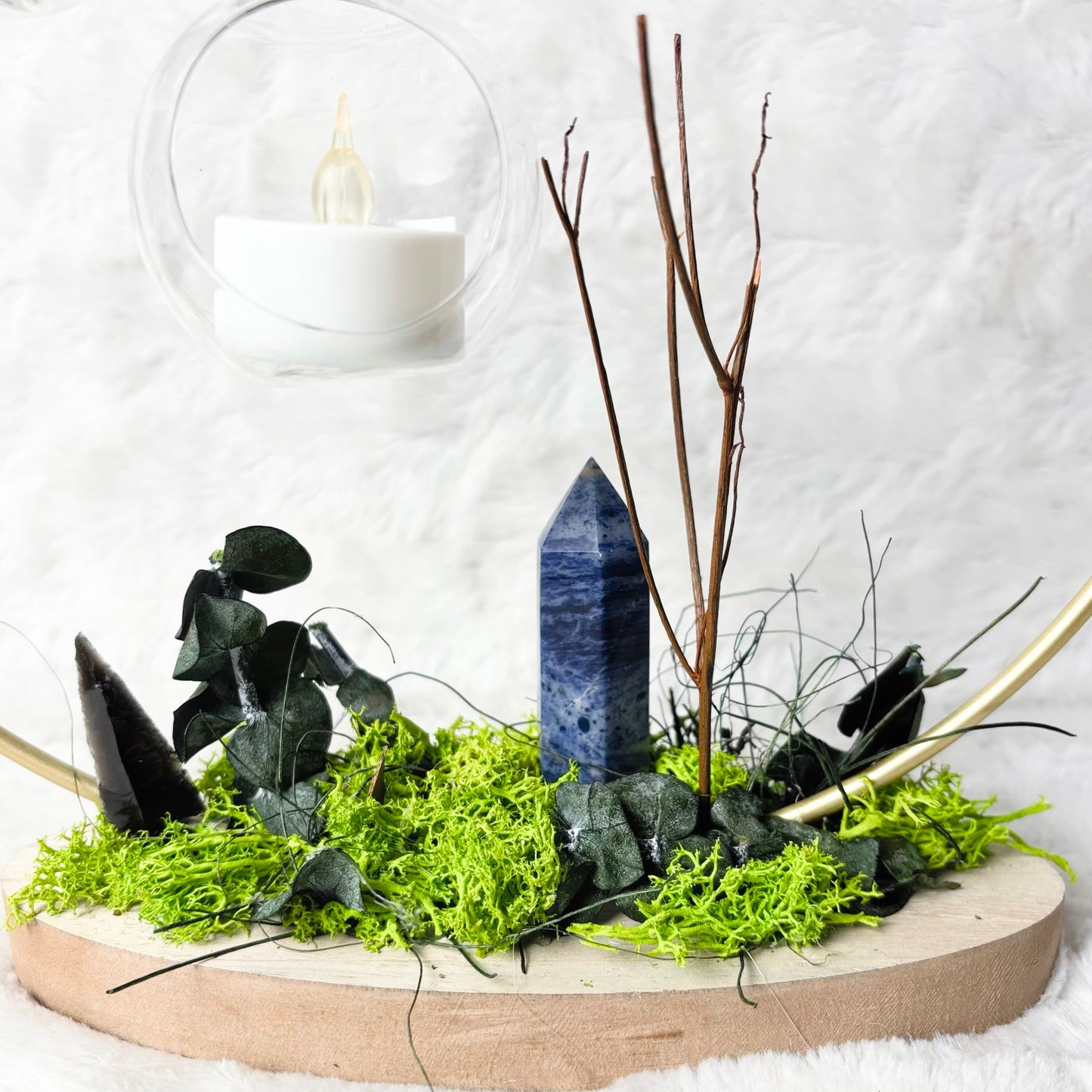 A decorative natural stone arrangement on a round wooden tray, featuring a blue lapis lazuli crystal point, black agate pieces, green moss, eucalyptus leaves, and bare twigs, set against a white faux fur background with hanging glass candle holders.